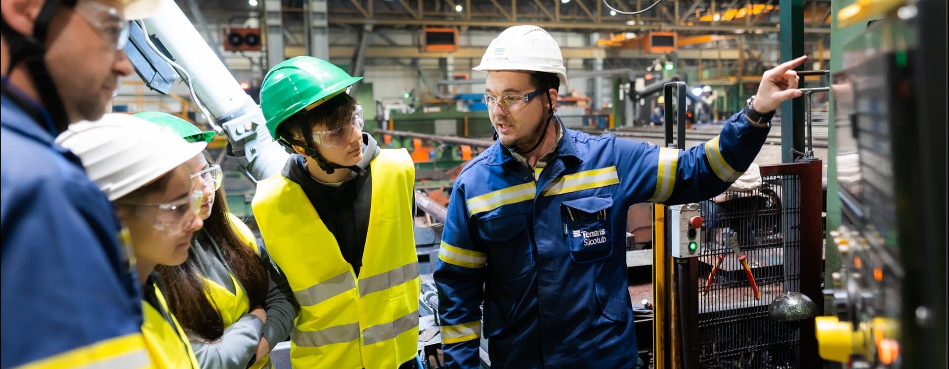 Elevi cu căști de protecție ascultând un inginer într-o hală industrială
