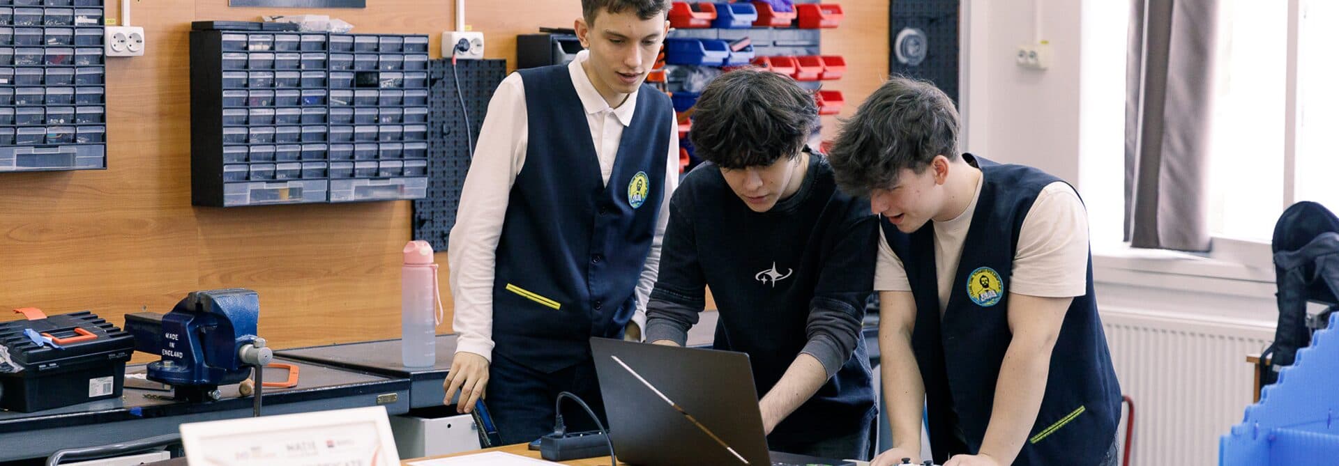 Trei elevi lucrează împreună la un laptop, într-un atelier