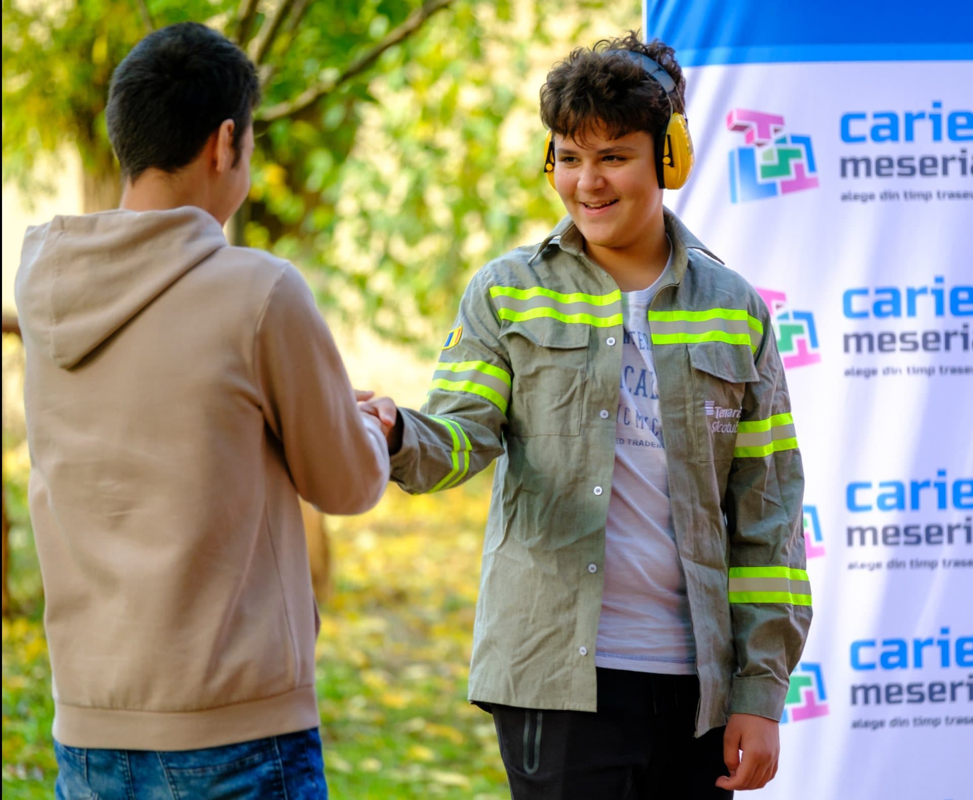 Fotografie: elev cu căști de protecție auditivă, dând mâna cu un adult, în fața unui banner Cariere Meseriașe